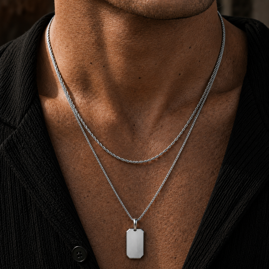 A close-up of a man's neck and chest wearing two layered silver chains. The shorter chain features a polished silver rectangular dog tag pendant with a brushed finish.