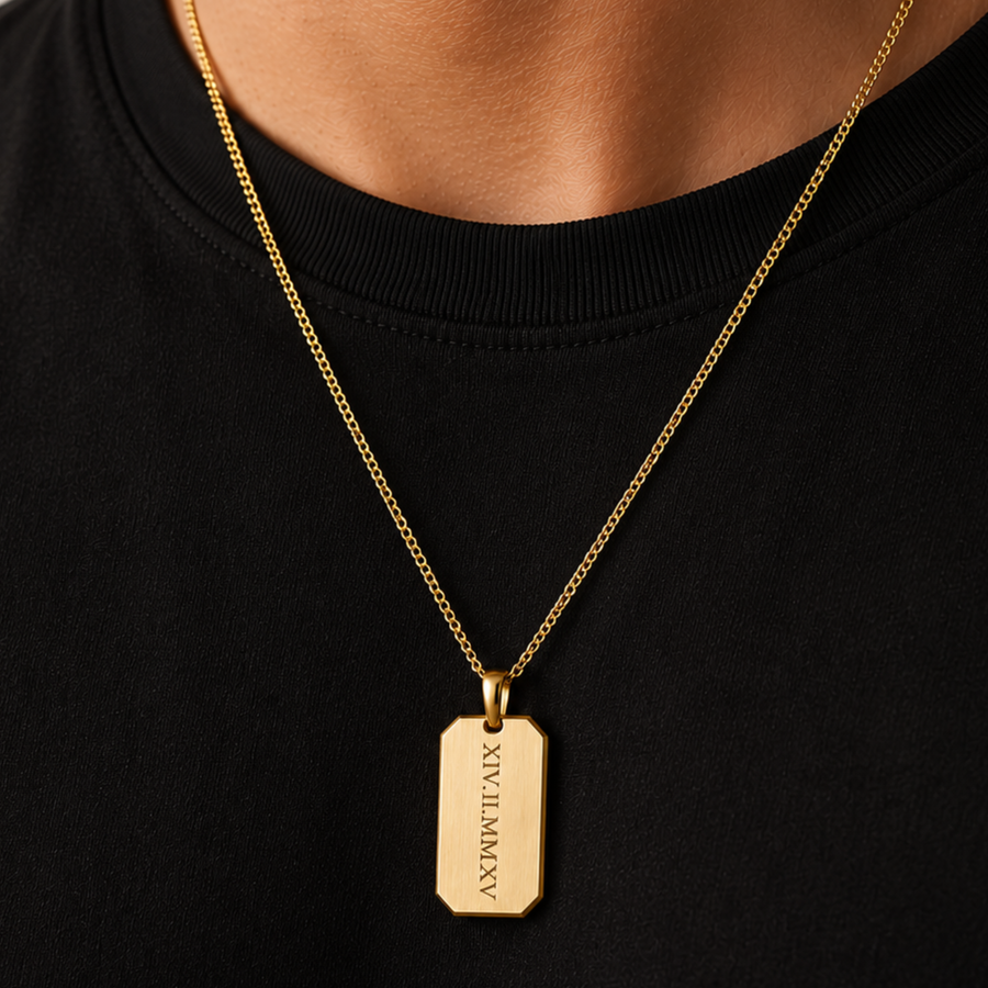 A gold-tone dog tag necklace with engraved Roman numerals, worn by a person in a black crewneck shirt.