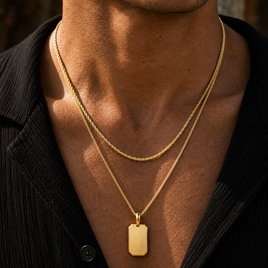 A close-up of a man's neck and chest wearing two layered gold-tone necklaces, including a flat rectangular dog tag pendant on a chain over a black shirt.