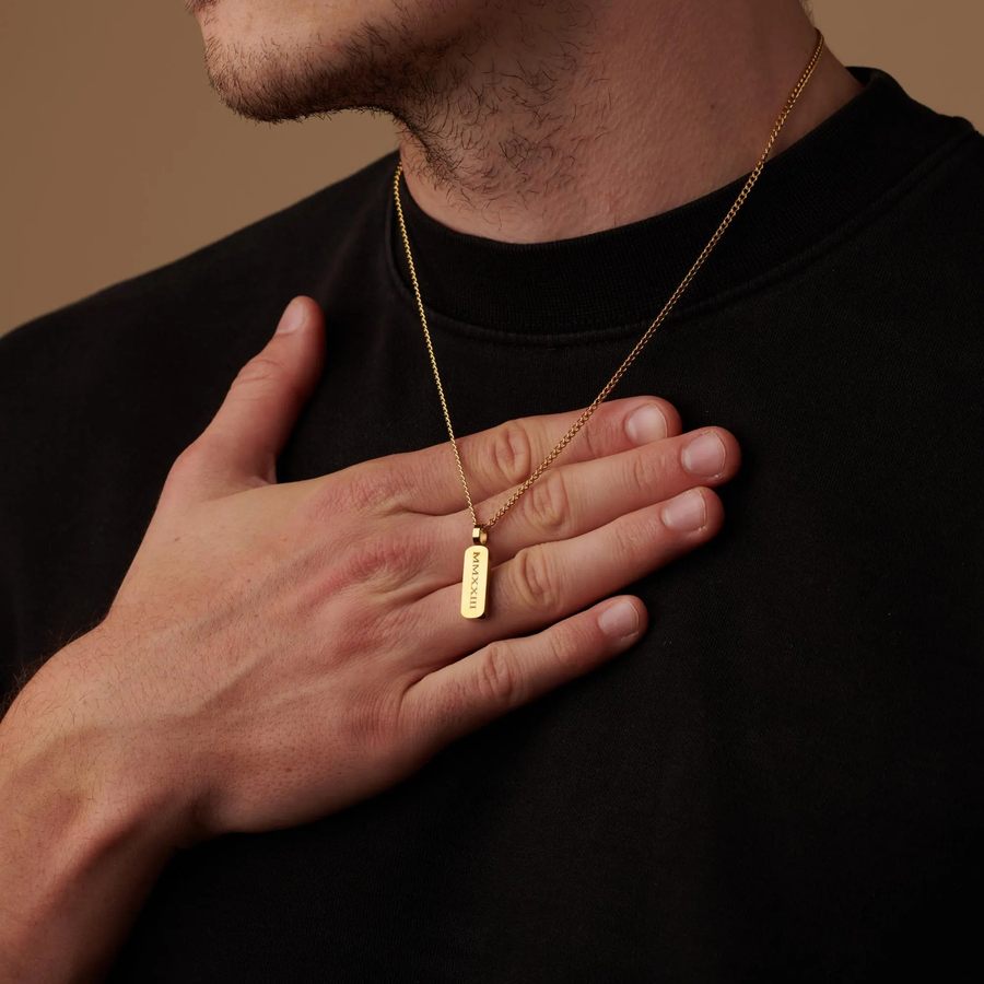A close-up of a man wearing a gold chain necklace with a small rectangular engraved pendant, resting his hand on his chest against a warm brown background.