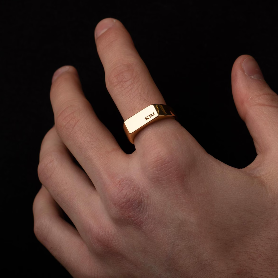 A close-up of a hand wearing a gold signet ring engraved with the name 'KIH' against a solid black background.