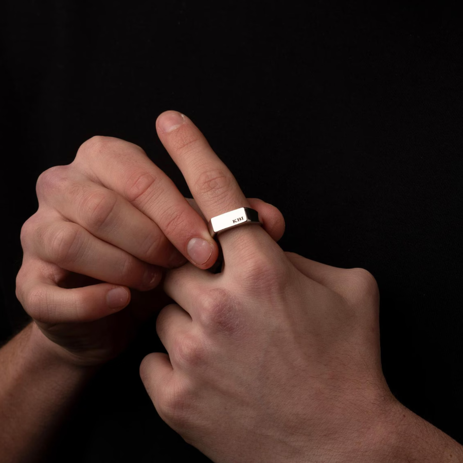 A close-up of a person's hands wearing a silver rectangular band ring engraved with 'KBI' on the side, set against a solid black background.