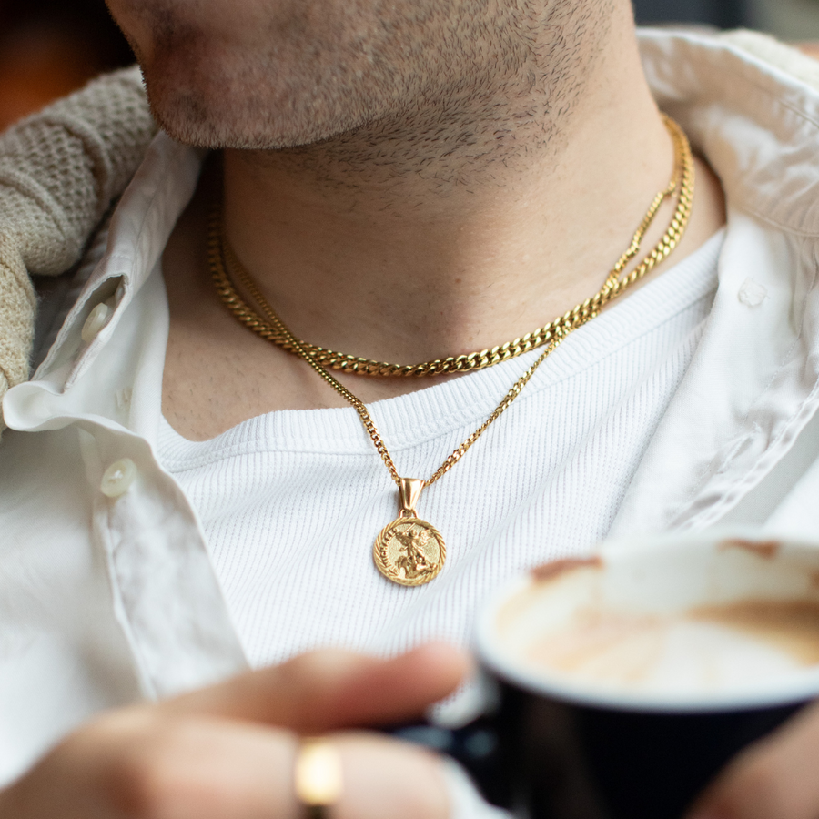 Person wearing a gold necklace with a pendant, holding a cup of coffee.