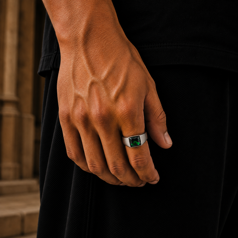 A close-up of a man's hand wearing a silver-toned ring with a square-cut green gemstone, set against a dark shirt and a blurred stone background.