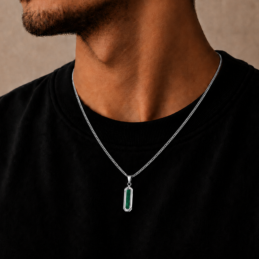 A close-up of a man wearing a silver chain necklace with a vertical rectangular pendant featuring a green inlay, set against a neutral beige background.