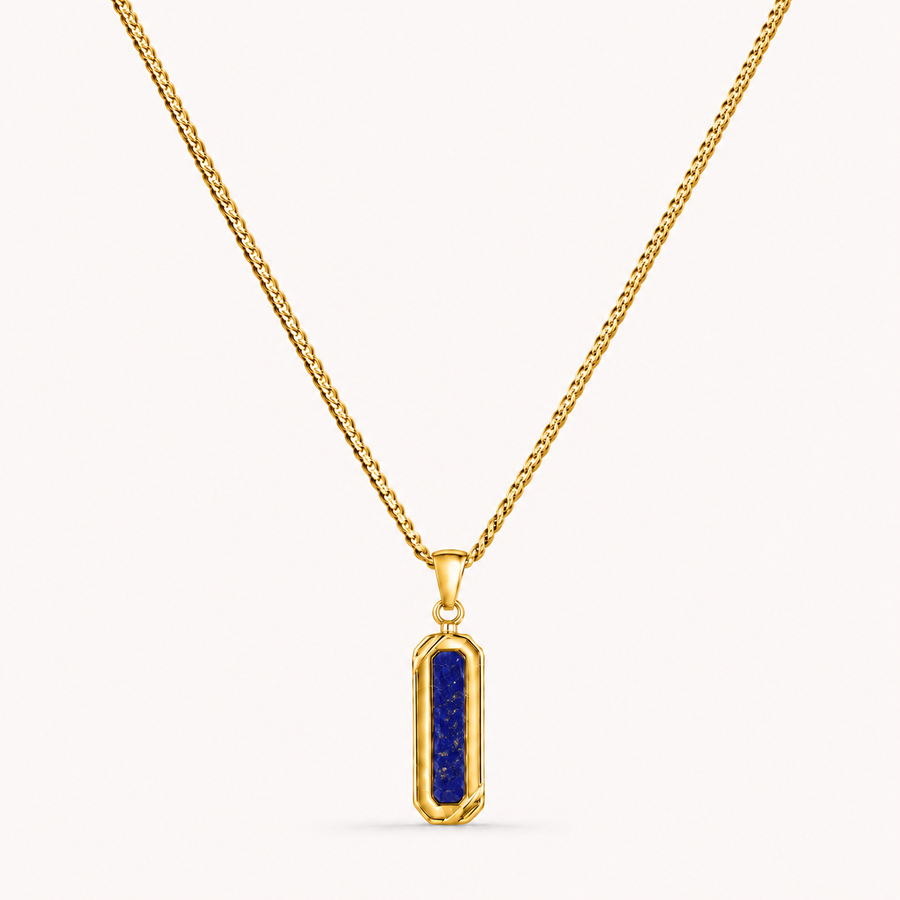 A gold chain necklace with a vertical rectangular pendant featuring a deep blue lapis lazuli crystal in a bezel setting, shown against a light off-white background.