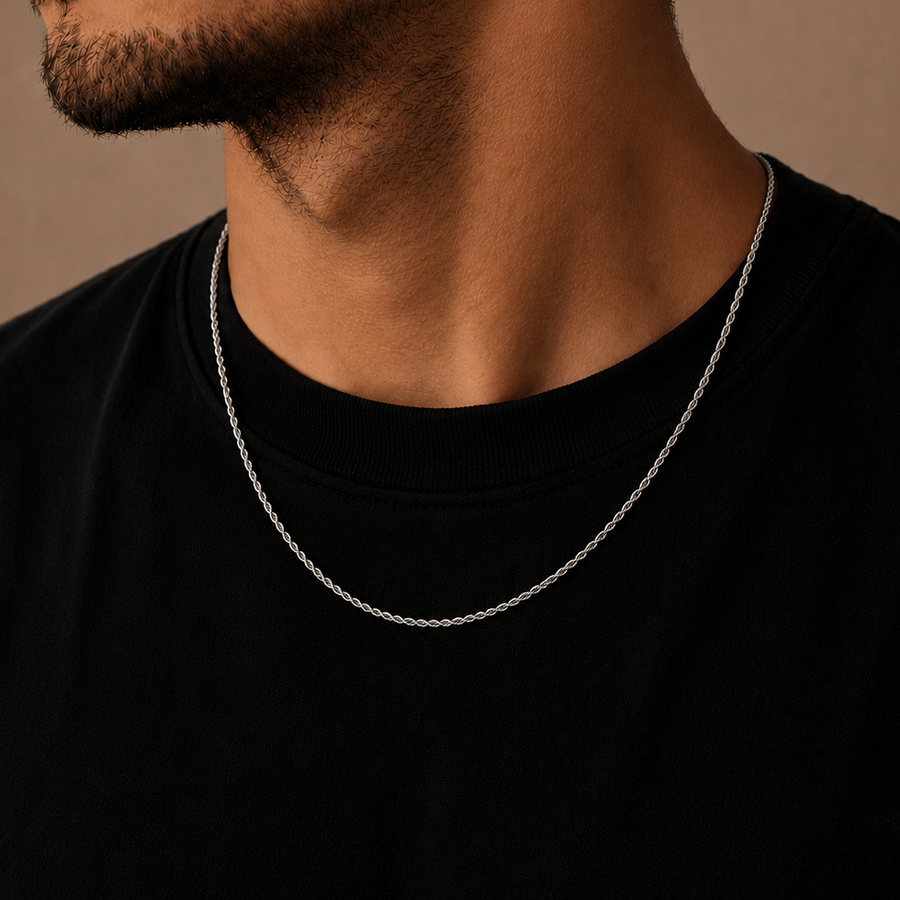 A close-up of a man's neck and lower face wearing a delicate silver chain necklace over a black t-shirt, set against a warm brown background.