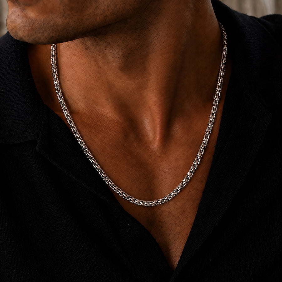 Close-up lifestyle image of a men’s polished silver wheat chain necklace, featuring a tightly woven interlocking link design worn around the neck with an open black collared shirt for a refined, understated luxury look