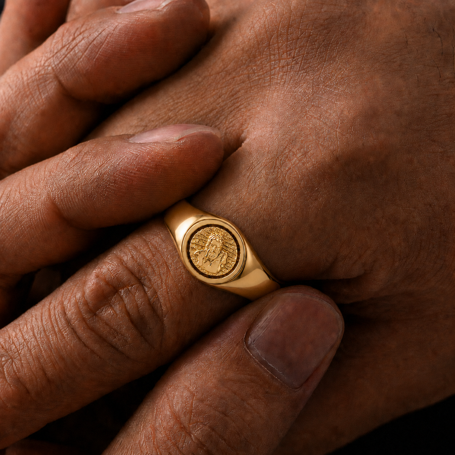 A close-up of a gold signet ring with an embossed coin-like face, worn on a finger of a person's hand.