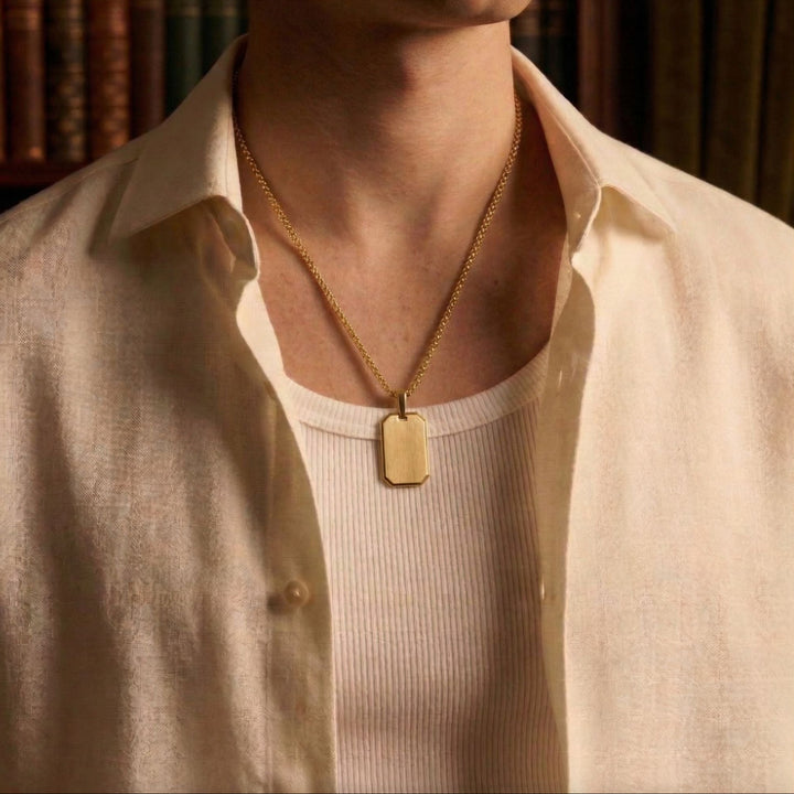 A close-up of a person wearing a gold chain necklace and a white shirt, with a small rectangular pendant hanging from the necklace.