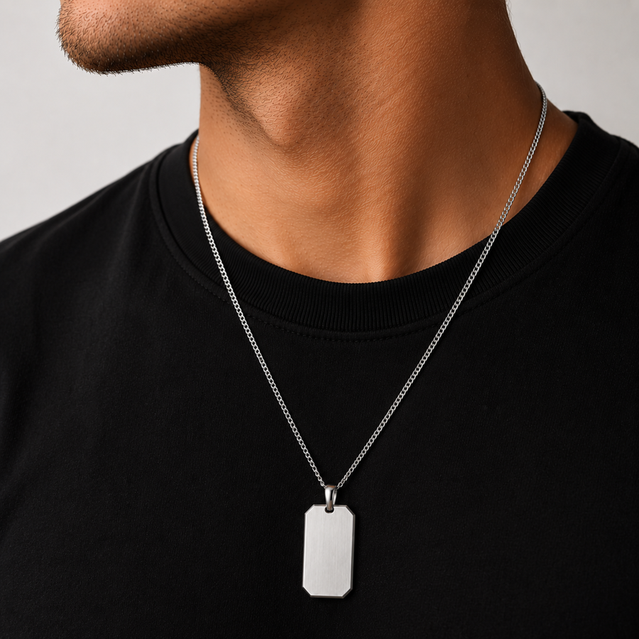 A close-up of a man's neck and chest wearing a silver chain necklace with a rectangular dog tag pendant, paired with a black crewneck shirt against a light gray background.