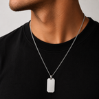A close-up of a man's neck and chest wearing a silver chain necklace with a rectangular dog tag pendant, paired with a black crewneck shirt against a light gray background.