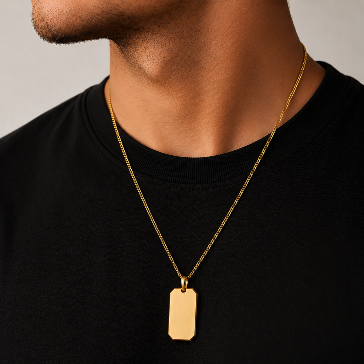 A close-up of a man's neck and chest wearing a gold chain necklace with a blank rectangular dog tag pendant, paired with a black crewneck shirt.