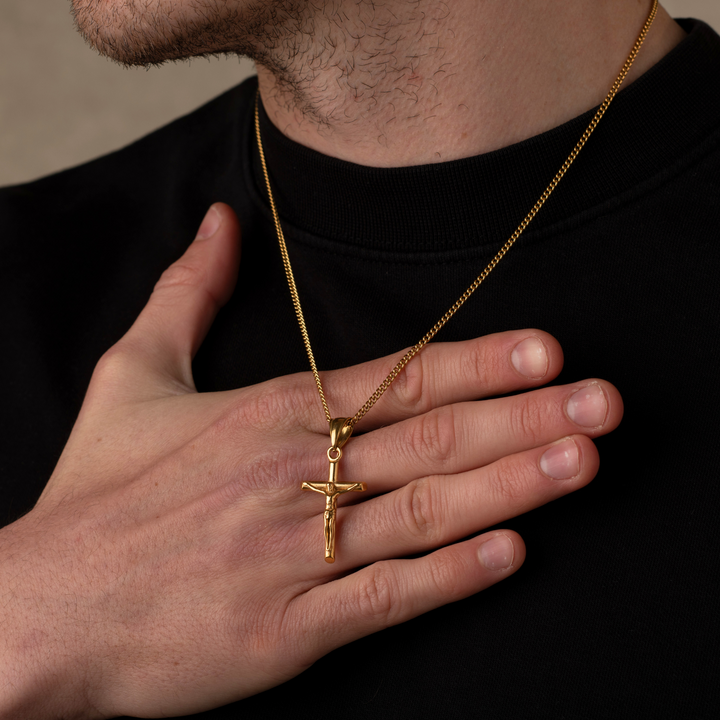 A close-up of a person wearing a gold crucifix pendant on a gold chain, with their hand resting on their chest over a black crewneck sweatshirt.
