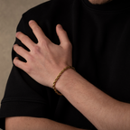 Hand wearing a gold bracelet on a black shirt against a neutral background