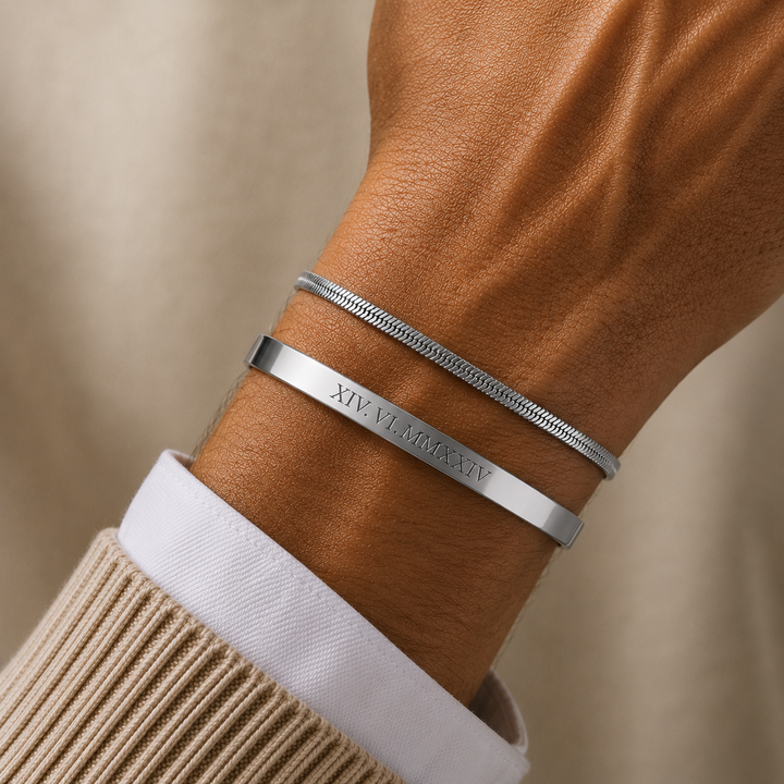 A close-up of a person's wrist wearing two silver bracelets, one smooth band engraved with 'XIV LXXXIV' and one textured snake-style bracelet, styled with a beige knit sweater and white dress shirt cuff against a soft beige background.