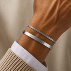 A close-up of a person's wrist wearing two silver bracelets, one smooth band engraved with 'XIV LXXXIV' and one textured snake-style bracelet, styled with a beige knit sweater and white dress shirt cuff against a soft beige background.