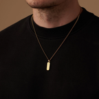 A close-up of a man wearing a black crewneck sweatshirt and a delicate gold chain necklace with a vertical rectangular nameplate pendant engraved with 'JAMES'.