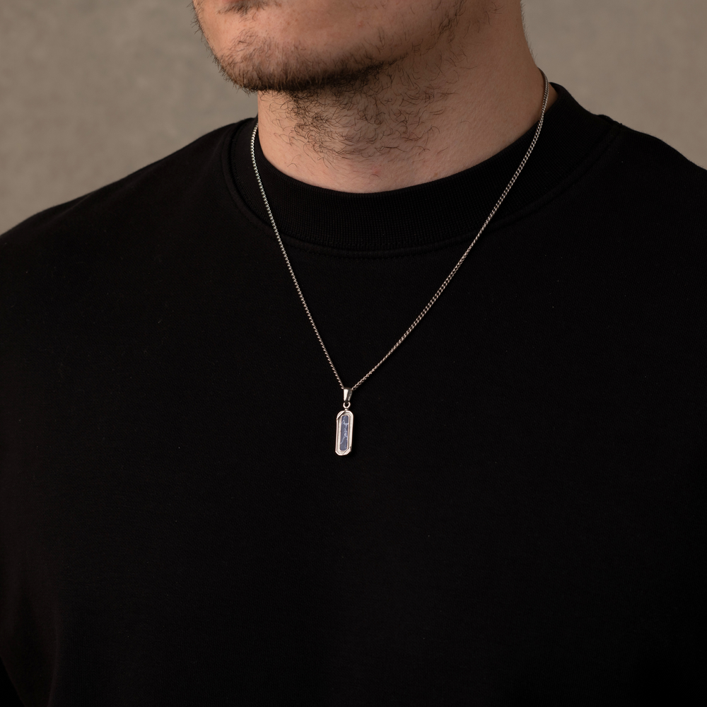 Man wearing a black shirt with a silver necklace featuring a rectangular pendant against a neutral background
