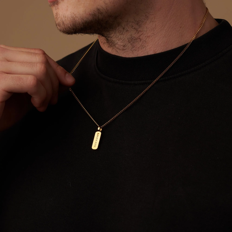 A close-up of a man wearing a gold chain necklace with a small rectangular engraved pendant, shown against a black shirt and warm beige background.