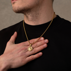 Man wearing a gold necklace with a pendant, holding it in his hand against a neutral background