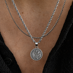 A close-up of a person wearing a silver double-strand rope chain necklace with a round Saint Patrick medallion pendant, styled over a black shirt.