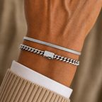 A close-up of a person's wrist wearing two silver bracelets: a thin snake-style bracelet and a thicker curb-link bracelet with a rectangular brand plaque.