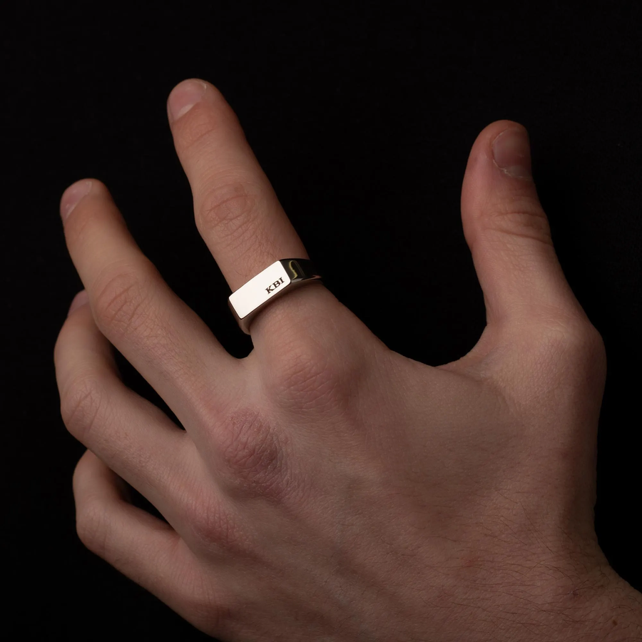 A silver rectangular band ring engraved with 'KBI' on the finger of a hand, shown against a solid black background.