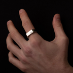A silver rectangular band ring engraved with 'KBI' on the finger of a hand, shown against a solid black background.