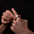 A close-up of a person's hands wearing a silver rectangular band ring engraved with 'KBI' on the side, set against a solid black background.