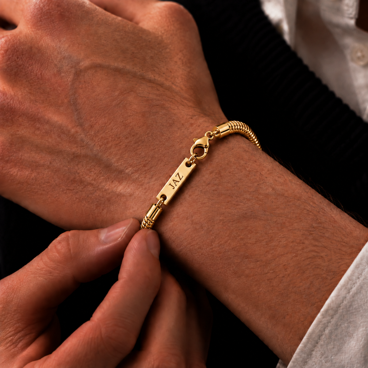 A close-up of a gold-plated personalized bracelet with a snake chain link and a flat bar engraved with 'JAZ' on the wrist.