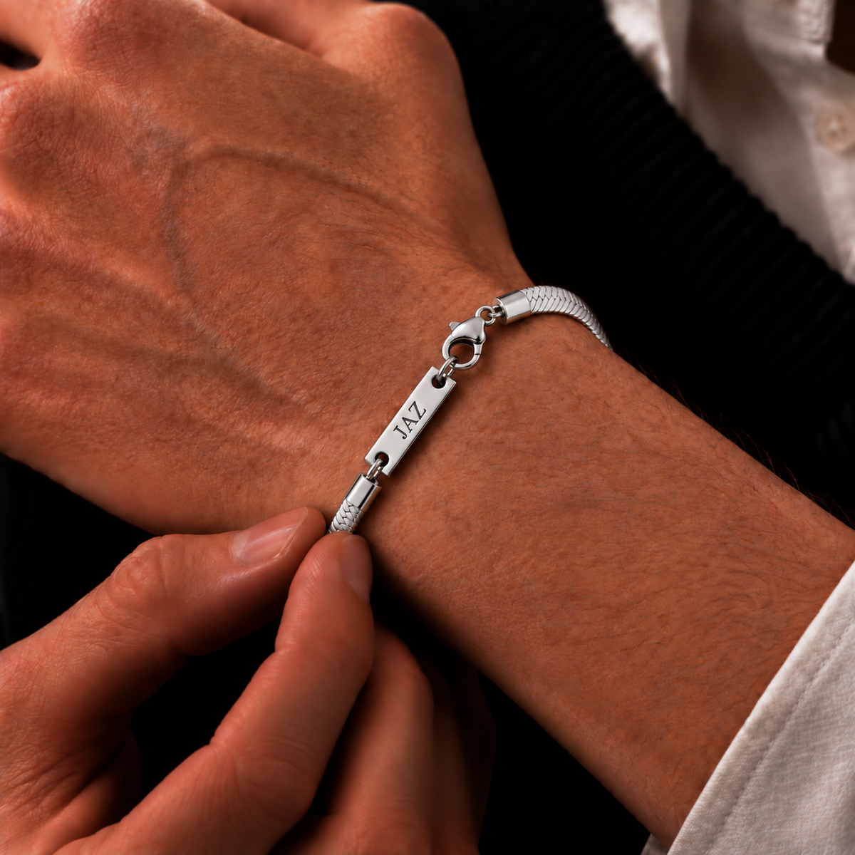 A close-up of a person's wrist wearing a silver nameplate bracelet with the name 'JAZZ' engraved on the central bar, with another hand adjusting the clasp.
