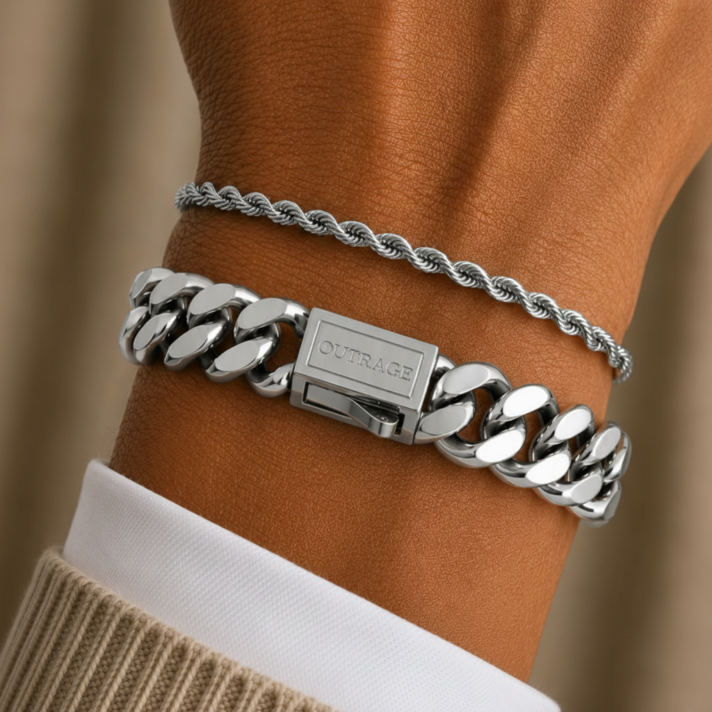 A close-up of a person's wrist wearing two silver bracelets: a thicker curb-link chain bracelet and a thinner twisted rope bracelet, layered over a white collared shirt and beige sweater.