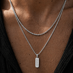 A close-up of a person's chest wearing two layered silver necklaces: one twisted chain and one longer chain with a rectangular pendant engraved with 'AMAXIV'.