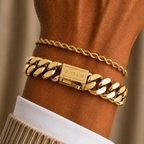 A close-up of a person's wrist wearing two gold bracelets: a thick curb-link chain bracelet with a rectangular clasp engraved with 'BURGA' and a thinner twisted rope chain bracelet, styled with a white dress shirt cuff and beige ribbed sweater.