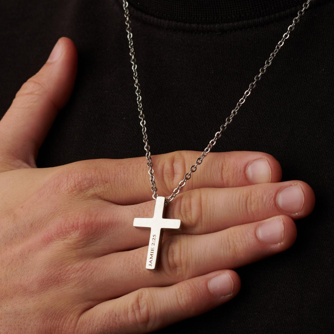 A silver cross necklace is worn around a person's neck, with the text "JAMIE 22.5" engraved on it.