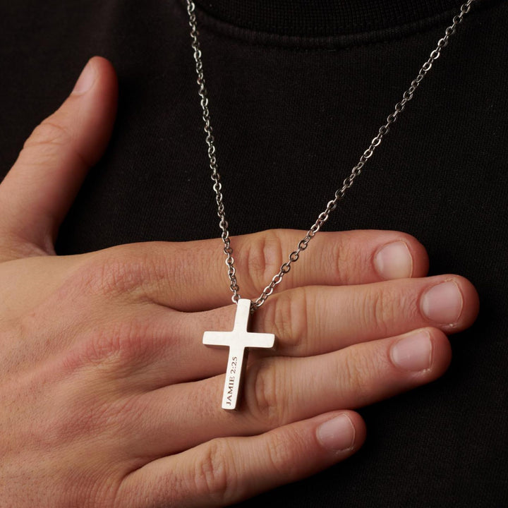 A silver cross necklace is worn around a person's neck, with the text "JAMIE 22.5" engraved on it.