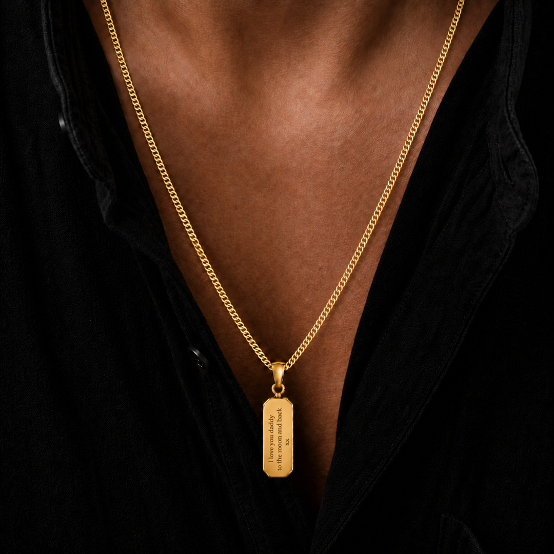 A close-up of a gold chain necklace with a rectangular gold pendant worn by a person in a black shirt. The pendant has engraved text on it.
