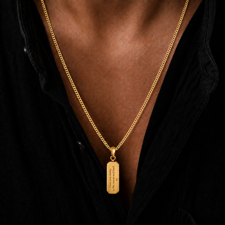 A close-up of a gold chain necklace with a rectangular gold pendant worn by a person in a black shirt. The pendant has engraved text on it.
