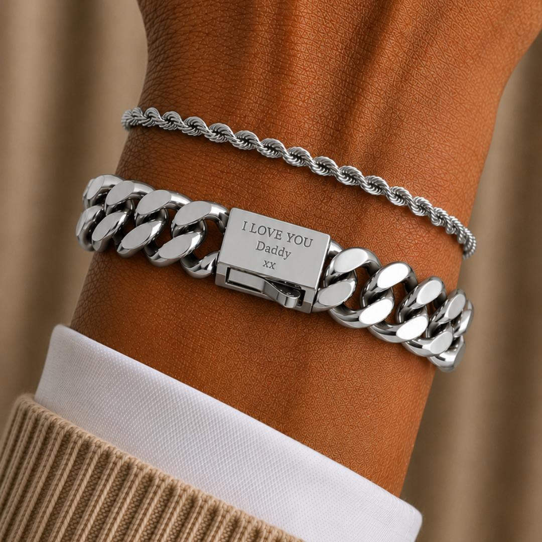 A close-up of a person's wrist wearing two silver bracelets: a thick curb chain with a rectangular plate engraved 'I LOVE YOU Daddy' and a thinner rope-style chain above it, shown with a beige knit sleeve and white shirt cuff against a warm brown background.