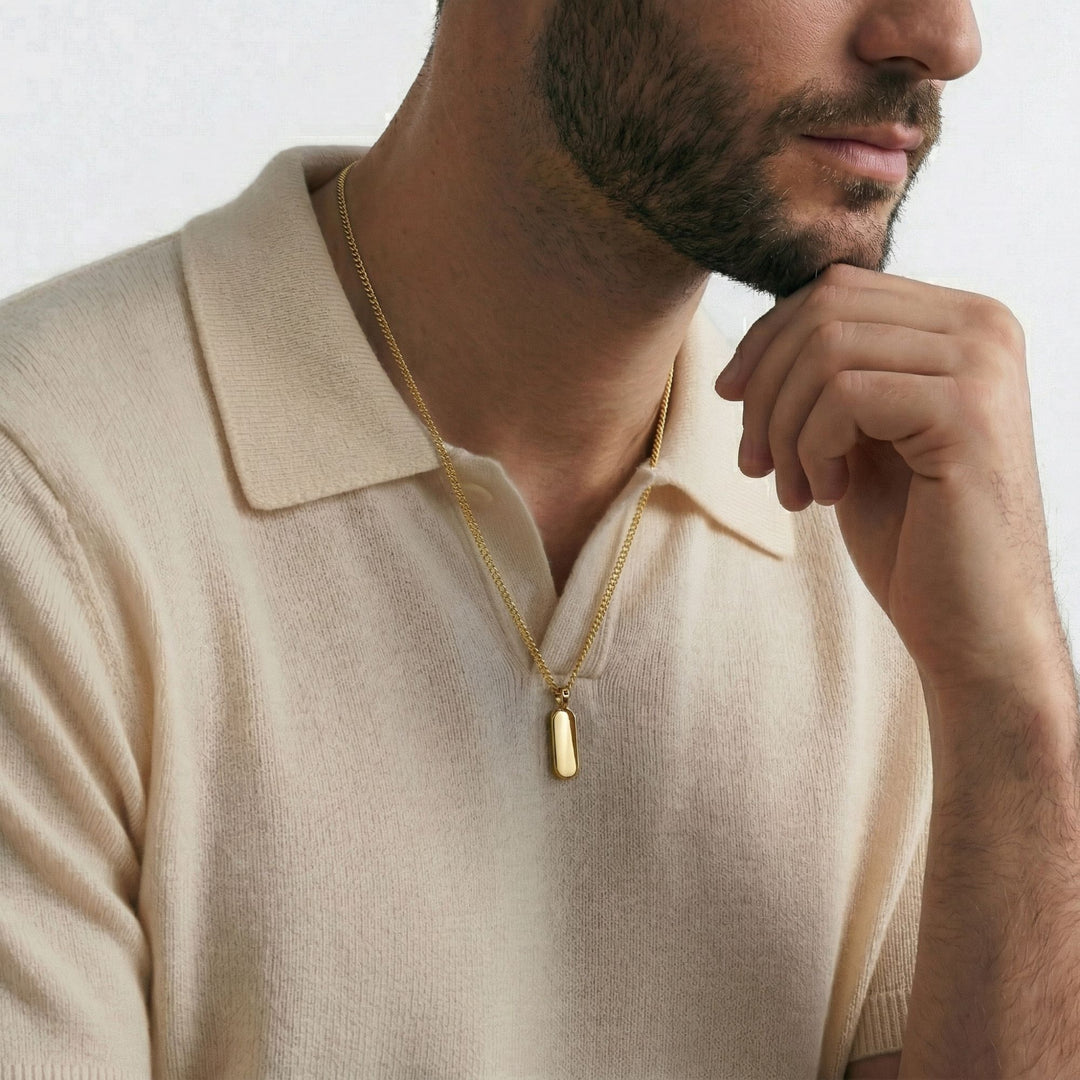 A close-up of a man wearing a gold necklace and a cream-colored polo shirt, with his hand resting on his chin.