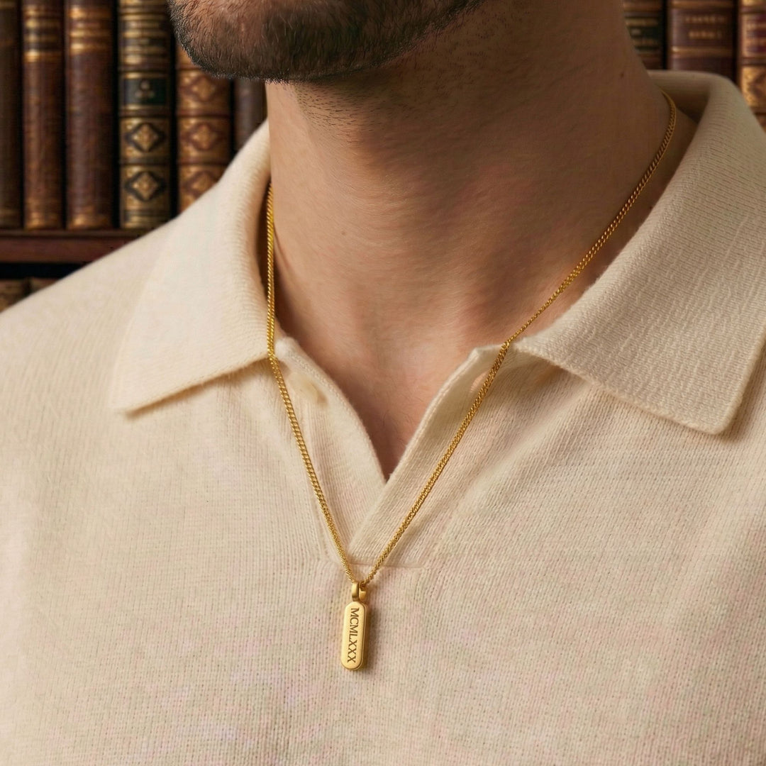 A close-up of a man wearing a gold necklace with a pendant, standing in front of a bookshelf.