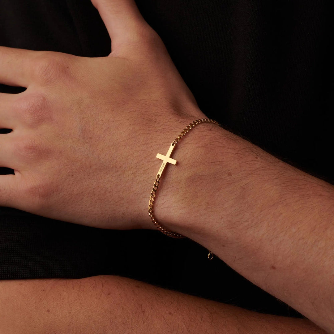 A gold cross bracelet is worn on a person's wrist.