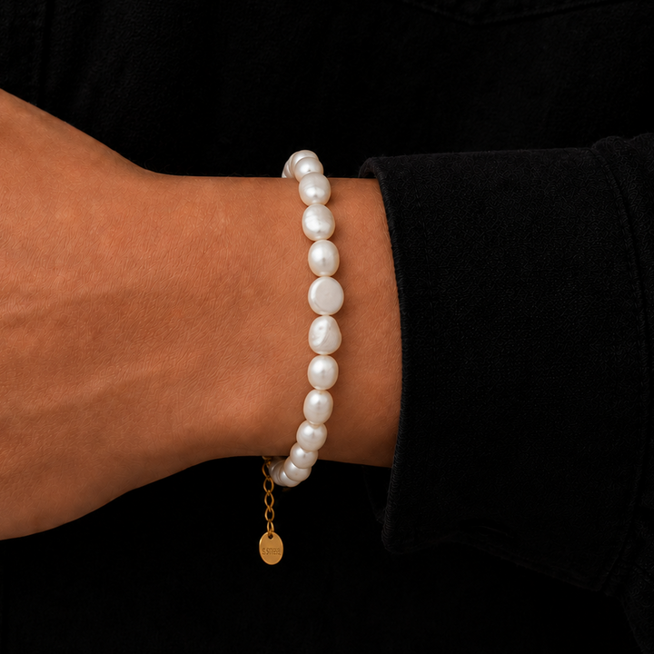 A close-up of a person's wrist wearing a beaded bracelet made of irregular white pearls with a small gold chain and round gold charm, set against a black shirt background.