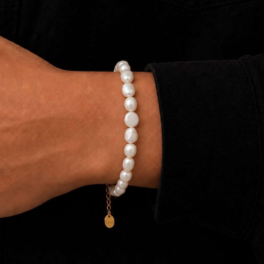 A close-up of a person's wrist wearing a beaded bracelet made of irregular white pearls with a small gold chain and round gold charm, set against a black shirt background.