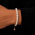 A close-up of a person's wrist wearing a beaded bracelet made of irregular white pearls with a small gold chain and round gold charm, set against a black shirt background.