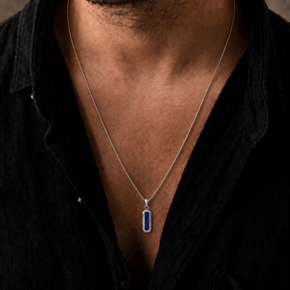 A close-up of a man wearing a silver chain necklace with a vertical rectangular blue pendant, styled with a black button-down shirt.