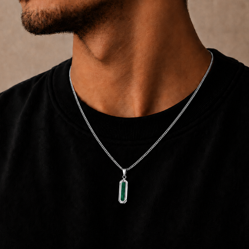 A close-up of a man wearing a silver chain necklace with a vertical rectangular pendant featuring a green inlay, set against a neutral beige background.