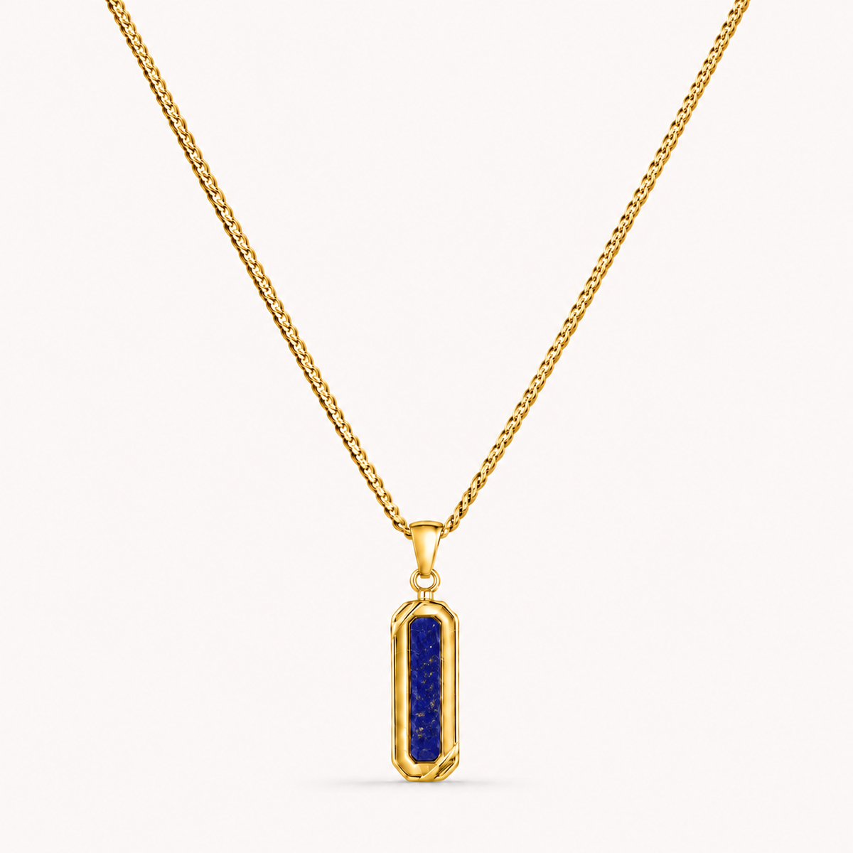 A gold chain necklace with a vertical rectangular pendant featuring a deep blue lapis lazuli crystal in a bezel setting, shown against a light off-white background.