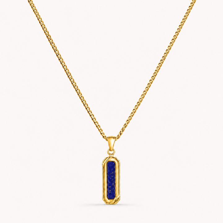 A gold chain necklace with a vertical rectangular pendant featuring a deep blue lapis lazuli crystal in a bezel setting, shown against a light off-white background.